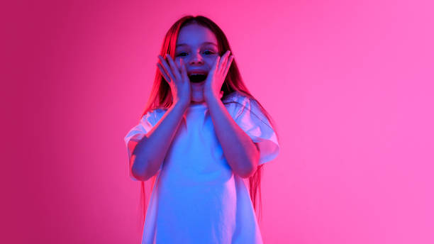 istock Girl with surprised expression holding hands on cheeks and looking excited in neon light against vivid gradient background. 2186753770