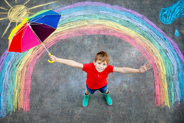 istock Happy kid boy in rubber boots with rainbow sun and clouds with rain drops painted with colorful chalks on ground or asphalt in summer. Creative leisure for children outdoors in summer 2186755010
