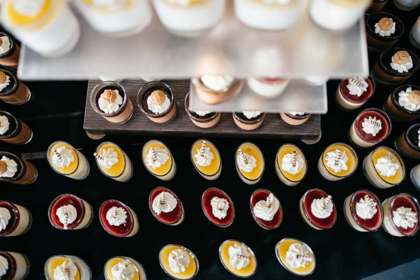 Elegant array of individual dessert shooters featuring layered puddings and mousses in clear shot glasses. Various flavors including vanilla panna cotta with mango coulis, chocolate mousse, and berry compote, each topped with fresh whipped cream and decorative wafer sticks. Perfect for catering, events, and luxury dessert presentations. Shallow depth of field with focus on creamy textures and vibrant toppings against dark background.