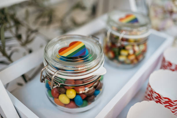 Close-up view of glass storage jars with clip closure containing colorful candies and decorated with rainbow pride heart stickers on top. The transparent containers are placed on a white surface, creating a modern and festive party setup. Perfect for celebrations, pride events, or candy bar arrangements.