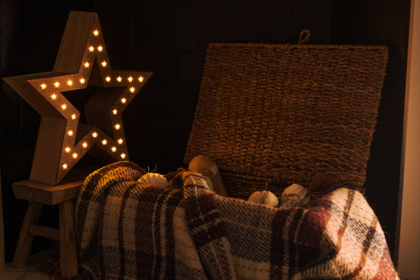 Christmas fire place cozy composition. Wicker basket chest with Christmas decorations and woolen blanket and wooden star lamp with soft cozy light. Dark cozy light