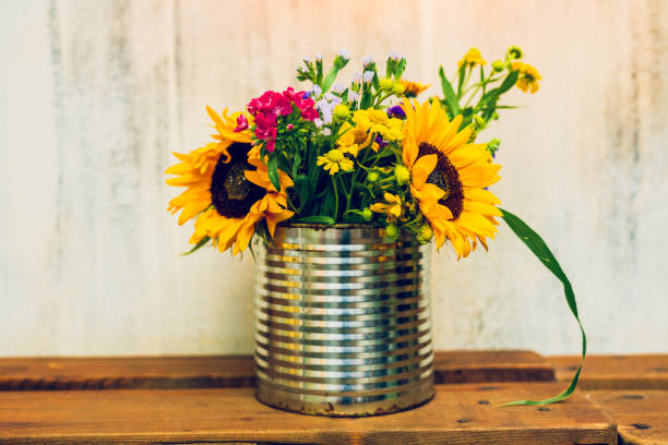 Lovingly arranged retro table decoration featuring a hand-picked wildflower bouquet with large sunflowers, elegantly displayed in a metal can on a wooden table, set against a vintage-style wall.