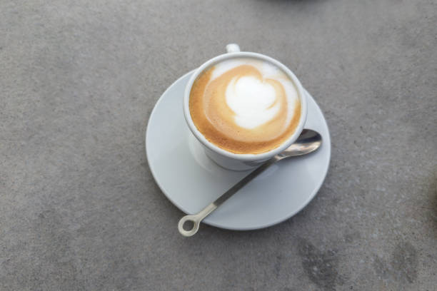Cappuccino coffee with drawing on a foam layer atop in white cup with a handle standing on saucer with spoon, top view on a gray table