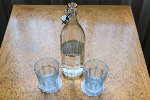 Glass bottle with ribbed outer surface and open reusable stopper half full with drink water and two empty faceted glasses stand on a small wooden table