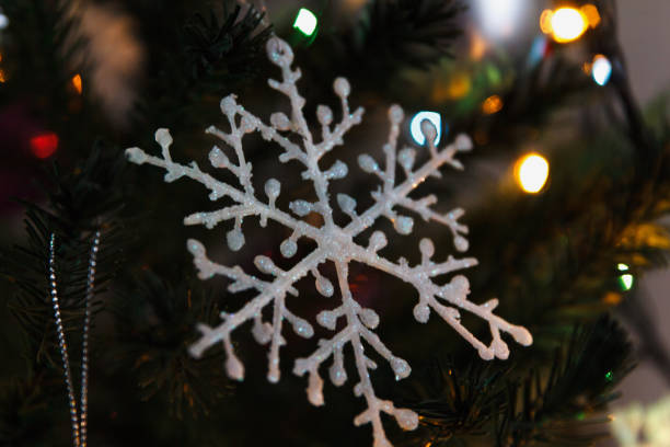 A hanging silver snowflake with a glowing bokeh background of warm lights and a blurred Christmas tree. Winter holiday concept.