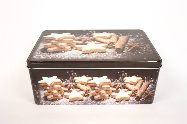 The image shows a rectangular metal tin with a decorative design featuring star-shaped cookies. The cookies are stacked in small piles and dusted with powdered sugar. Cinnamon sticks and star anise are also part of the design, adding a festive and cozy feel to the tin. The tin is likely used for storing cookies or other baked goods, making it both functional and visually appealing.