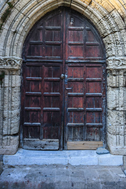 Two weathered wooden arched doors and one closed rusted wrought iron window on bricks stone wall and tiled stone floor
