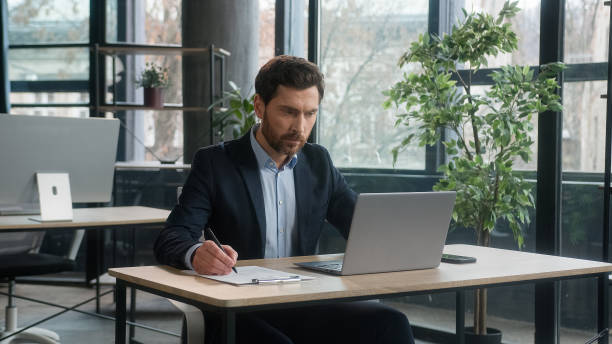 istock Focused adult businessman work on project in office using laptop writing notes caucasian man business leader professional employee analyze information on internet write data planning with computer app 2186820035