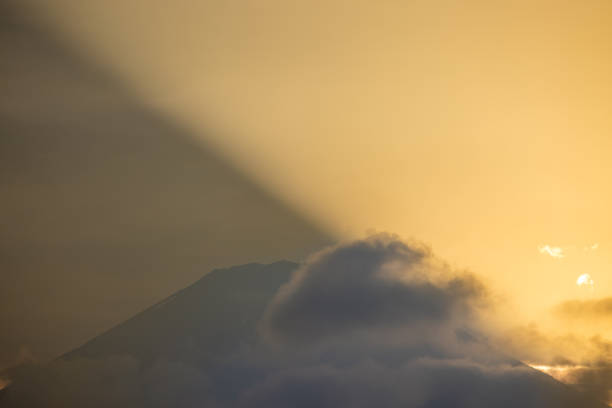 A breathtaking view of Mount Fuji partially obscured by clouds during a stunning sunrise. The golden hues of the sky and the dramatic beam of sunlight create a captivating and ethereal atmosphere. This iconic Japanese landmark is beautifully highlighted in a moment of natural splendor, perfect for showcasing the majesty of Japan’s landscapes.
