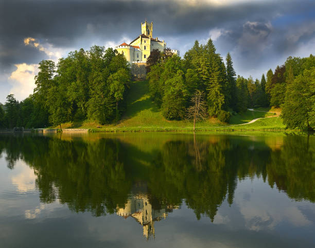 A view from Bled Castle of Lake Bled on a sunny fall day, with the iconic Church of the Assumption of the Blessed Virgin Mary on Bled Island. Surrounded by vibrant autumn foliage, the serene lake reflects scattered clouds against a blue sky. The church, a 17th-century Baroque masterpiece, remains one of Slovenia’s most revered landmarks. Visitors often take traditional pletna boats to the island to admire the church's stunning architecture and peaceful atmosphere.