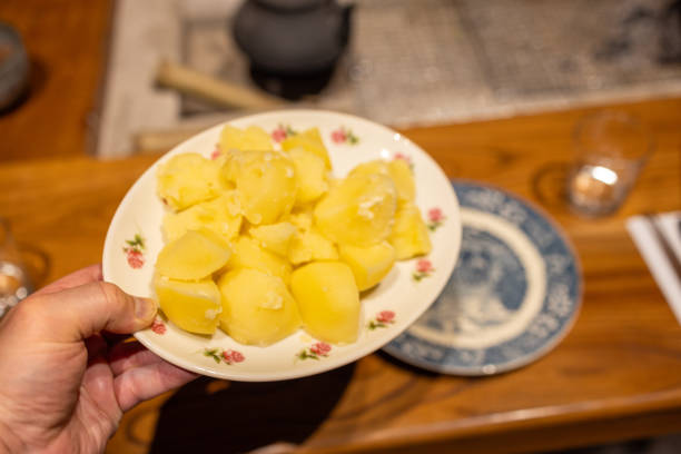 A hand presenting a floral-patterned plate filled with freshly boiled potatoes. The soft, golden potatoes are steaming hot and ready to serve, offering a comforting and wholesome addition to any meal. The rustic table setting in the background, complete with traditional dishware, adds warmth and charm to this culinary moment. Perfect for showcasing homemade cooking, comfort food, or simple yet nourishing meal ideas.