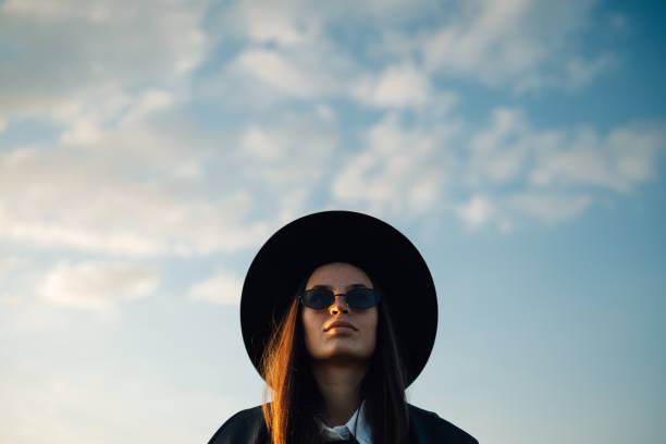 Stylish woman wearing a wide brimmed hat and sunglasses poses confidently against a backdrop of a cloudy sky, creating a fashionable and evocative scene