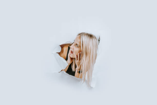 Valmiera, Latvia - November 24, 2024 - A blonde woman peeks through torn white paper with an expressive and curious look, creating a modern, creative, and minimalistic portrait.