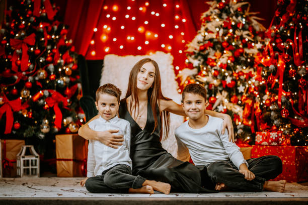 A young woman sits with two boys in a festive Christmas setting, surrounded by decorated trees, glowing lights, and wrapped gifts, sharing smiles.