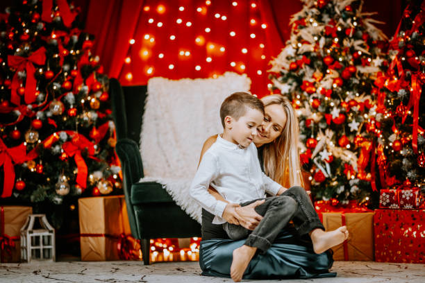 A loving mother embraces her son in a warm, festive Christmas setting with glowing lights, decorated trees, and wrapped gifts in the background.