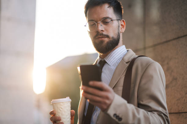 Businessman using smartphone and holding coffee outside office building at sunset