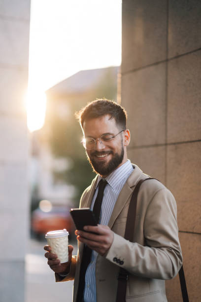 istock Smiling businessman using smartphone and holding coffee outside office building at sunset 2186861495