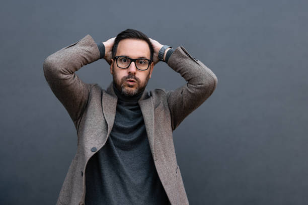 The image features a man with glasses holding his head with both hands, expressing surprise or realization. His eyes are wide open, conveying intense and immediate emotion, while the neutral gray background brings focus to his striking gesture and facial expression.