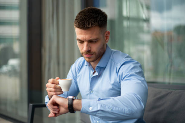 istock Young Businessman Sitting in Cafe, Checking Time While Holding Coffee, in Front of Modern Business Building 2186867659