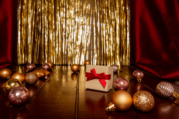 Festive scene featuring a wrapped gift box with a red ribbon, surrounded by shiny golden and red baubles on a polished wooden surface, with a golden curtain backdrop. Christmas postcard