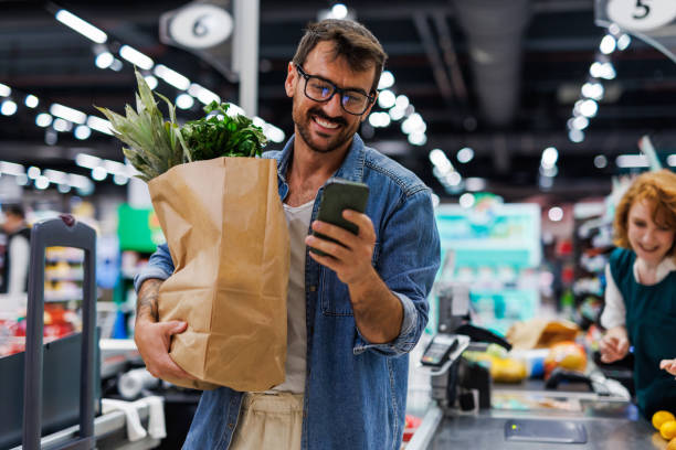 smiling customer using smartphone while holding grocery bag at checkout - kassa bildbanksfoton och bilder