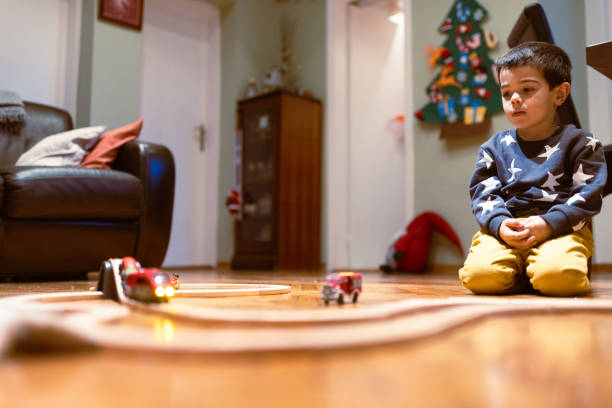 A young child sitting on the floor near a Christmas tree is intently watching a toy train set in a cozy living room, capturing the magic and wonder of the holiday season.