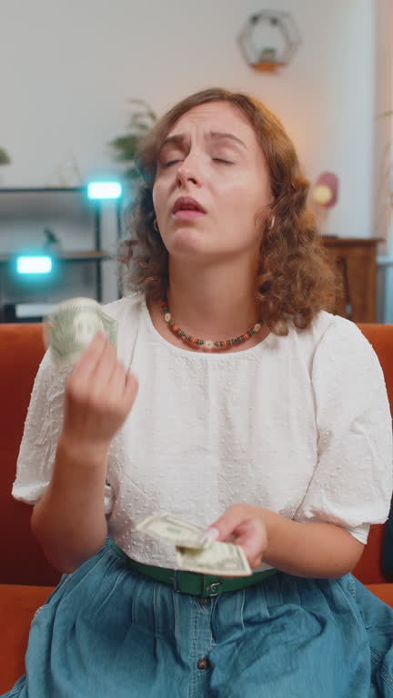 Upset young woman sitting on sofa counting money, having not enough, worried about finances, debts