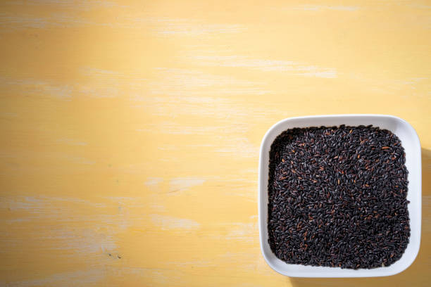 A bowl of black raw rice is placed on a yellow table.The raw rice is spread out in the bowl, copy-space.
