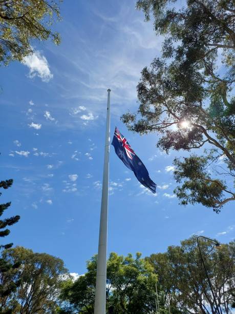 istock Australian flag at half-mast on a tall flagpole under a bright blue sky, framed by sunlit trees, creating a solemn and respectful atmosphere. 2186893155