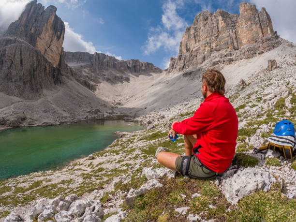 A hiker in a red jacket sits on rocky terrain overlooking a serene green alpine lake, surrounded by towering cliffs and a bright blue sky.