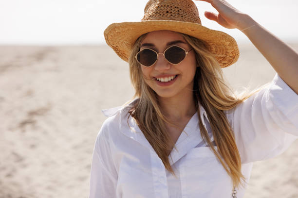 Close-up portrait of beautiful young woman with straw hat and sunglasses standing on sandy beach on windy spring day