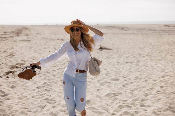 Beautiful young woman wearing jeans, white shirt and straw hat happily running along sandy beach