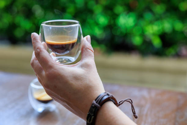 Hand holding a cup of espresso in transparent glass cup at wooden table outdoors, morning ritual