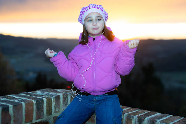 A cute little girl listens to music with her earphones, sitting on a low wall against a dramatic sunset sky in Cereseto, overlooking the hills of Monferrato, a UNESCO World Heritage Site.