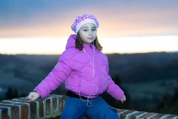A cute little girl listens to music with her earphones, sitting on a low wall against a dramatic sunset sky in Cereseto, overlooking the hills of Monferrato, a UNESCO World Heritage Site.