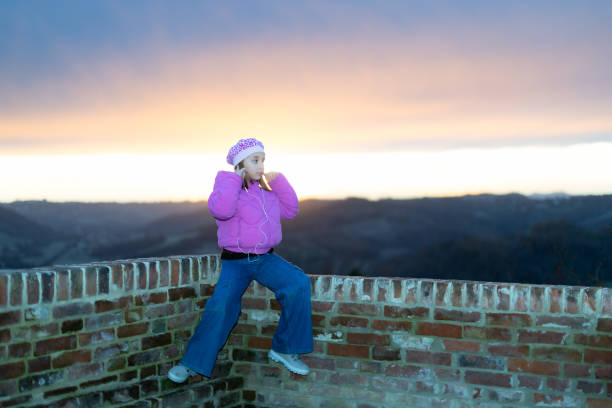 A cute little girl listens to music with her earphones, sitting on a low wall against a dramatic sunset sky in Cereseto, overlooking the hills of Monferrato, a UNESCO World Heritage Site.