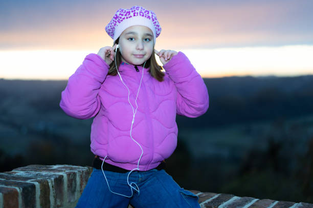 A cute little girl listens to music with her earphones, sitting on a low wall against a dramatic sunset sky in Cereseto, overlooking the hills of Monferrato, a UNESCO World Heritage Site.