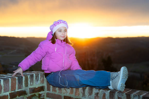 A cute little girl listens to music with her earphones, sitting on a low wall against a dramatic sunset sky in Cereseto, overlooking the hills of Monferrato, a UNESCO World Heritage Site.