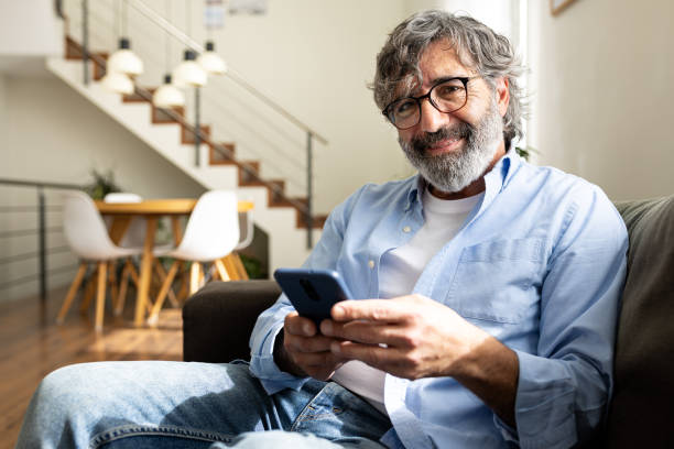 istock Mature man relaxing at home, sitting on the couch using phone looking at camera. 2186940408