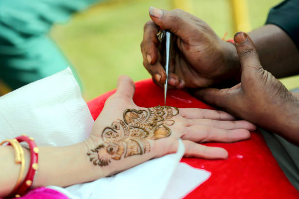 An artist applying intricate henna (mehndi) designs to a person's hand, creating beautiful patterns in a traditional and artistic style. The scene reflects a festive or ceremonial moment.