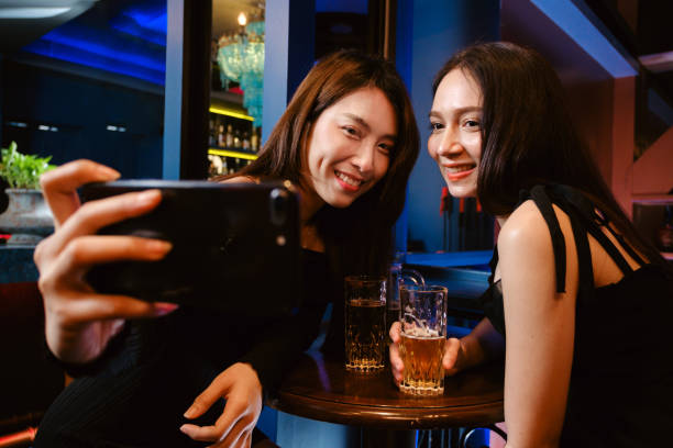 Two women smiling while taking a selfie at a bar, with drinks in front of them, enjoying a lively night out in a colorful ambiance.