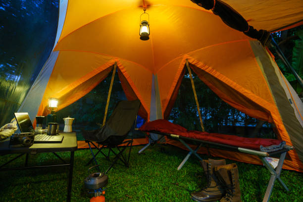 Camping tent, folding bed inside the tent at night in the forest, gas lantern and coffee set, notebook on a  camping table, and outdoor ideas.