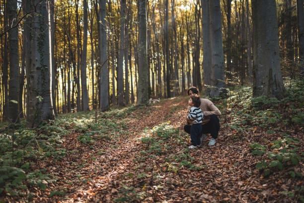 A mother and child share a tender moment in a serene forest, surrounded by autumn leaves and tall trees, highlighting family connection and nature's beauty.