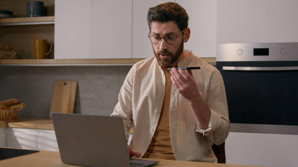 istock Multitasking Caucasian man working with laptop recording dictating voice message voicemail with phone at home kitchen. Male freelancer businessman with computer communicating distant with mobile app 2186979542