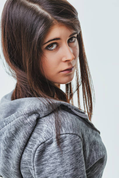 Young woman wearing a gray hoodie turns her head to look over her shoulder, her intense gaze conveying a mix of emotions