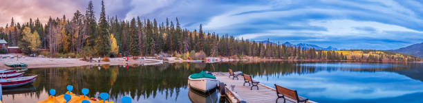 A serene view of Pyramid Lake in Jasper National Park, Canada. Calm waters reflect the vibrant autumn forest, docked boats, and distant mountains under a dramatic evening sky.