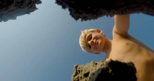 A young boy curiously peers down from a height at a beautiful rocky beach beneath a clear blue sky