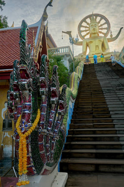 The image captures the majestic Buddha statue at Wat Plai Laem in Koh Samui, Thailand. The colorful staircase adorned with Naga sculptures leads worshippers to the impressive figure, creating a peaceful and spiritual atmosphere.