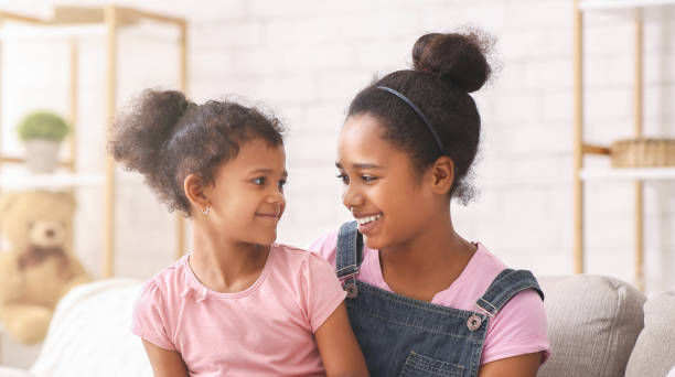 Happy african american sisters smiling to each other, sitting at home, panorama