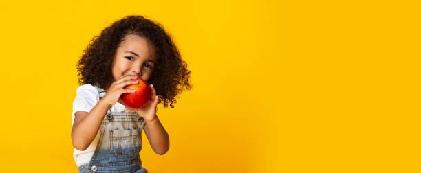 istock Cute afro girl biting red apple, yellow background 2187053805